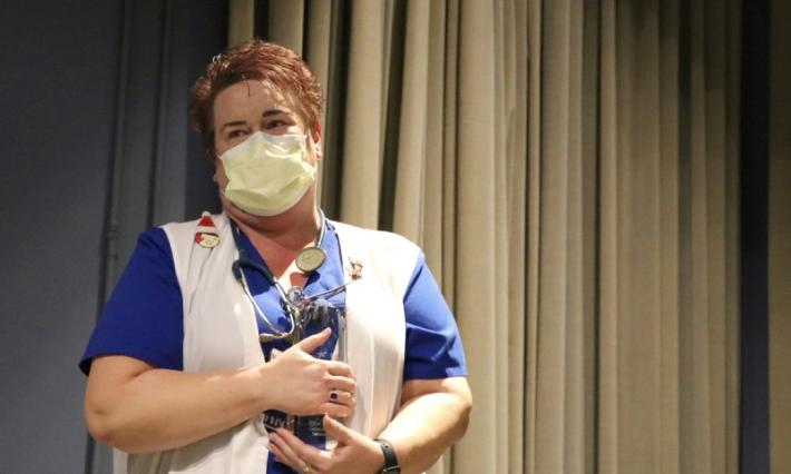 Woman in a facemask and vest, with a stethoscope around her neck, holds a glass award to her chest