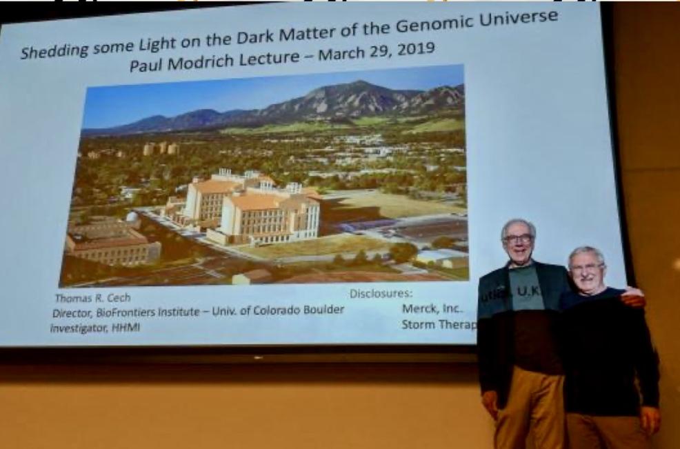 Thomas Cech and Paul Modrich stand in front of a large screen with the projection of an aerial view of BioFrontiers Institute at the University of Colorado Boulder and the title of Cech's presentation.