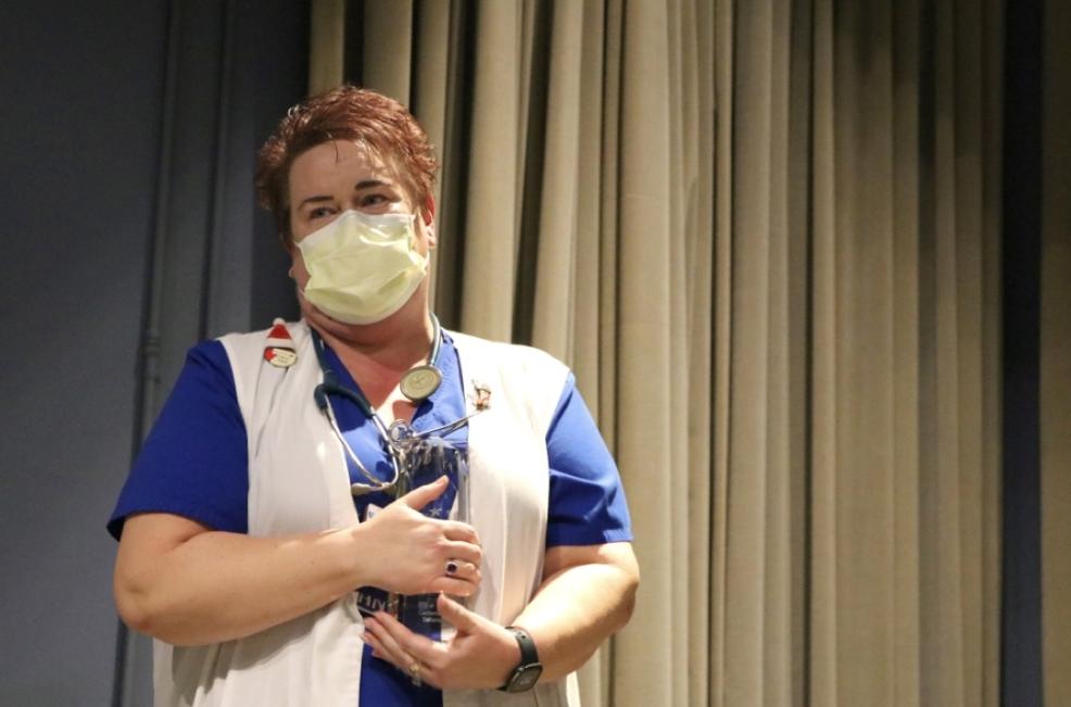 Woman in a facemask and vest, with a stethoscope around her neck, holds a glass award to her chest