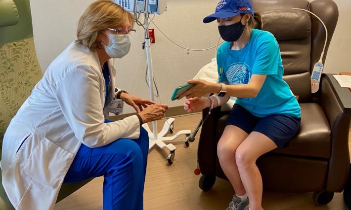 At left, clinical trial nurse Leighanne Hartman has a white labcoat and mask on. Elle Charnisky in a baseball cap and t-shirt is showing her photos while receiving a chemotherapy infusion.