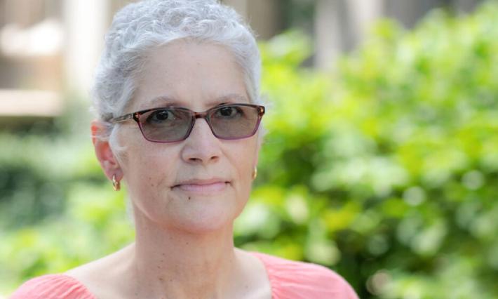 Sharlene Mitchell is outdoors in this headshot wearing sunglasses, short white/gray hair, and a pink top. There is a large bush in the background.