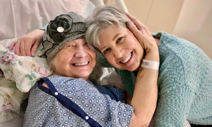 Kathy Jennings hugs her friend Rebecca Gordon, who is wearing a hospital gown and a hat and sitting on a hospital bed.