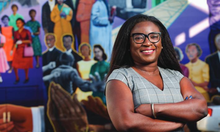 Tomi Akinyemiju smiles with arms folded in front of a colorful mural