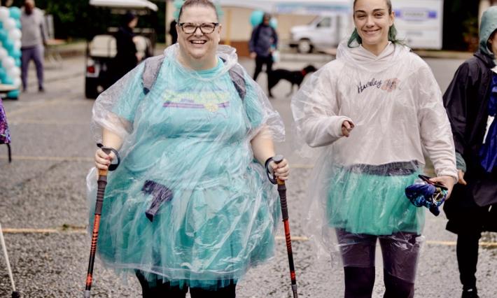 A woman dressed all in teal with a clear rain poncho smiles. She walks with two canes and with a smiling female teenager beside her.