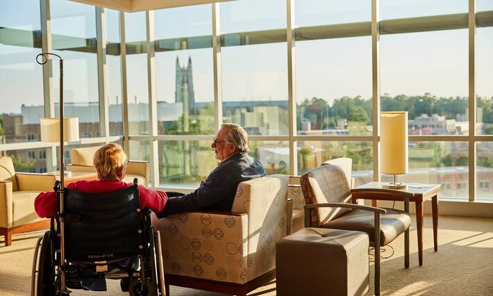 A woman in a wheelchair with her hsband in front of a window overlooking DCI buildings and greenery.