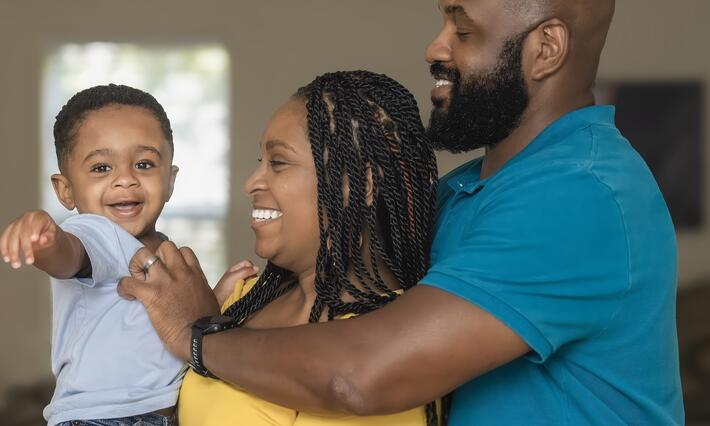 Two parents, one holding a toddler, smiling together as the toddler reaches toward the camera.