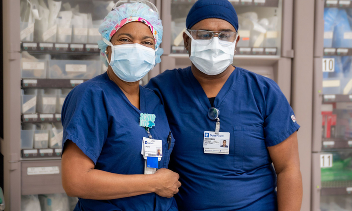 Two Duke Health team members dressed in blue scrubs, surgical masks, and caps stand together in the OR.
