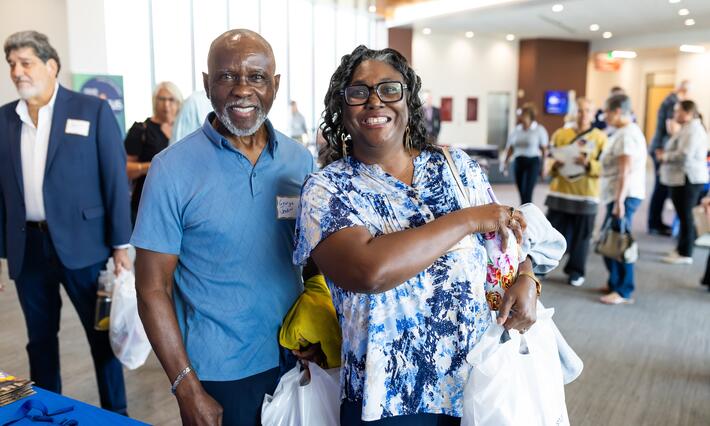 Black man with gre beard and black woman holding white bag with Duke CAncer Institute logo. They are standing in a large gallery filled with people