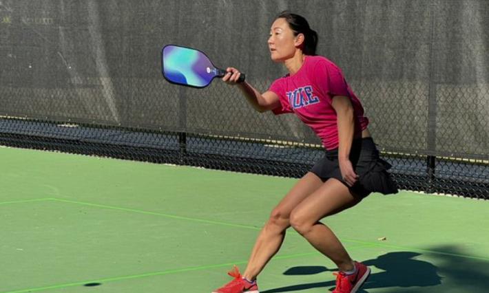 woman in shorts and pink t-shirt with Duke written on it leans back on the green court; holding up her racquet to hit the ball 