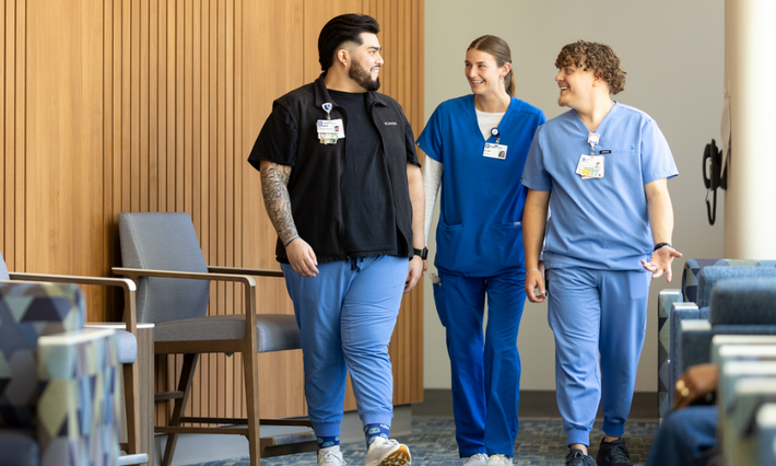 Three Duke Health team members wearing scrubs of varying shades of blue walk together through the hospital hallway.