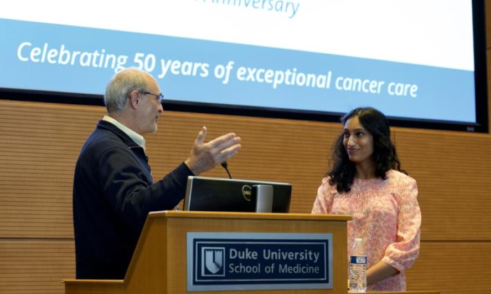 Man stands at a Duke University School of Medicine podium facing woman smiling. "Duke Cancer Institute 50th Anniversary: Celebrating 50 years of exceptional cancer care" is projected on the screen behind them.