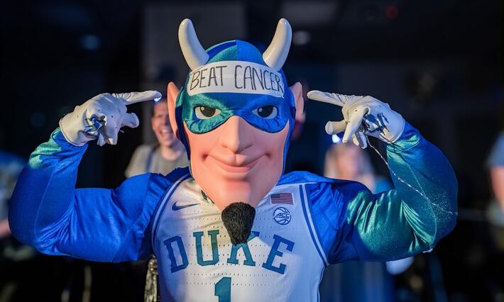 The Duke Blue Devil Mascot wears a Duke basketball jersey and a cape and points to a sign on his forehead that reads "Beat Cancer"