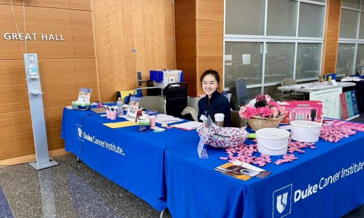 a woman smiles behind a table draped in blue with the words Duke Cancer Institute. The table is covered with pink decorations, papers, and other items