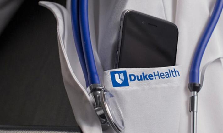 A close-up of a doctor's white coat, which says "Duke Health" on the pocket. An iPhone sits inside the pocket and a stethoscope hangs down in frame. 