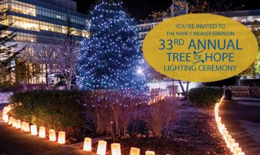 tree lit with blue lights at night and luminaries along the garden paths on either side with illuminated buildings in the background, plus the words "33rd Annual Tree of Hope Lighting Ceremony" in a yellow circle
