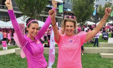 Two women dressed in pink raise their hands together and smile while standing on the grass