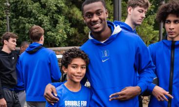 Outdoors a man in a Duke sweatshirt with arm around a child in Dribble for Victory t-shirt holding a basketball. Four other team members in the background