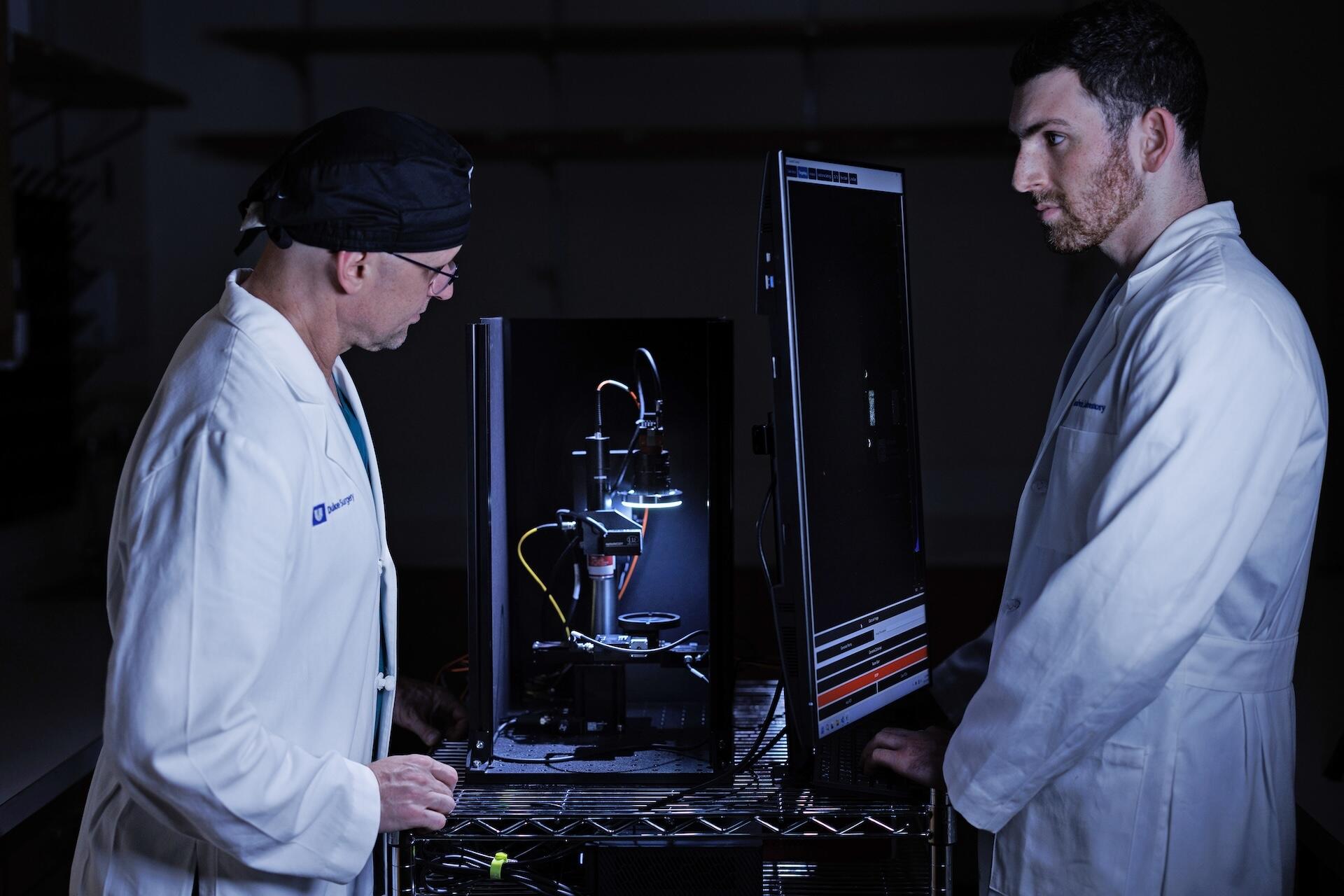 Two men in white coats standing together working with a microscope, screens, and other elements of the Prostate Tumor ID device