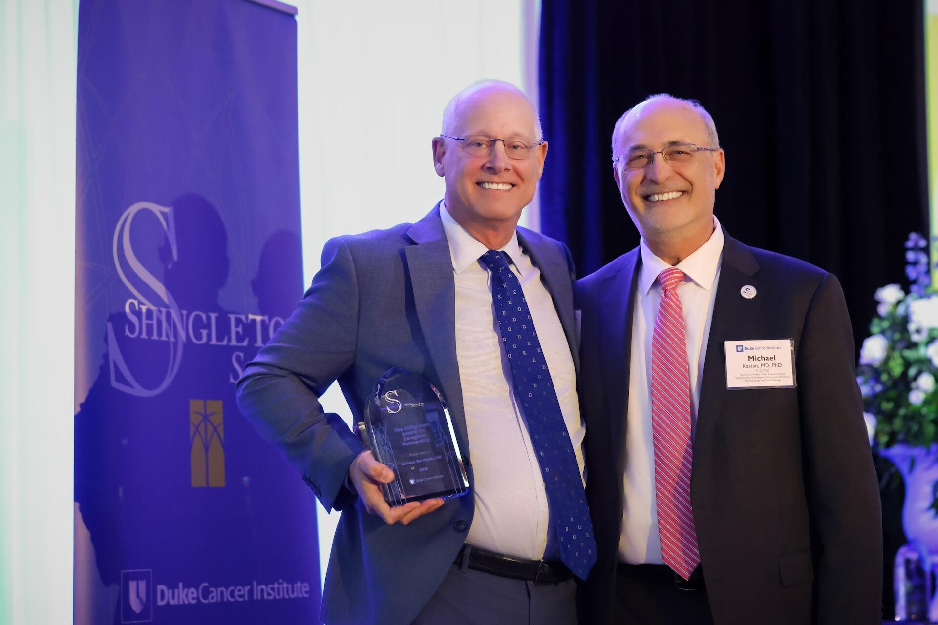 1.	Two men in suits smiling together, one holds a crystal Shingleton trophy.