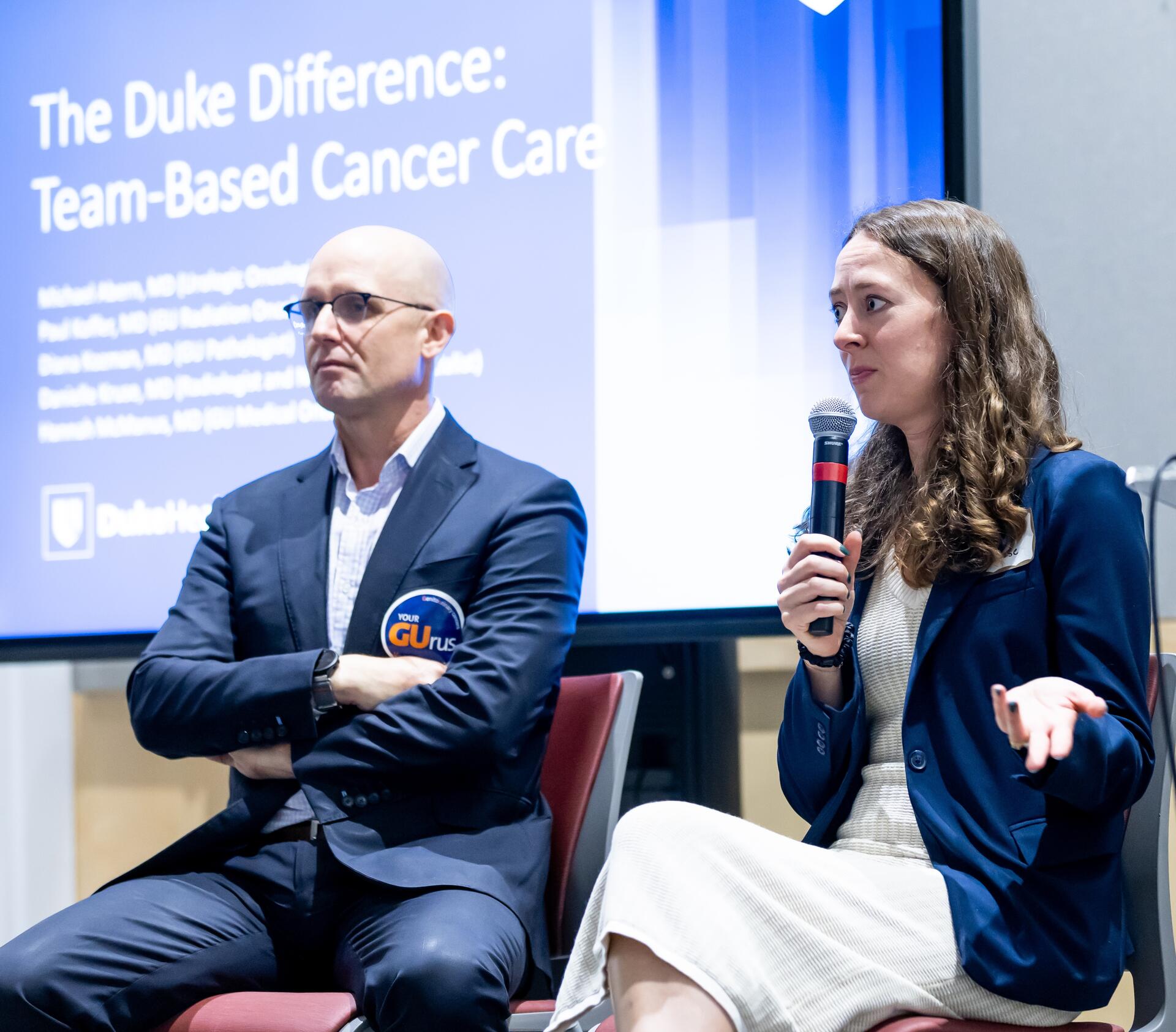 A Man and Woman in business dress on stage. Behind them is a slide with title "The Duke Difference: Team-Based Cancer Care."
