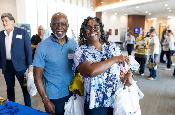 Black man with gre beard and black woman holding white bag with Duke CAncer Institute logo. They are standing in a large gallery filled with people