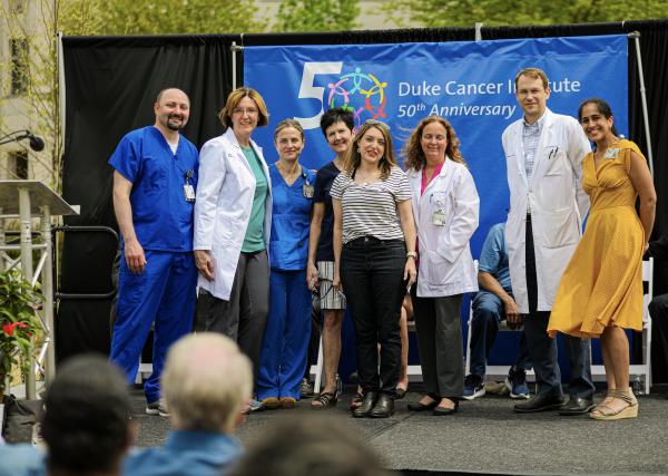 Seven DCI Team Members in scrubs and labcoats stand with Elle Charnisky on stage in front of a DCI 50th Anniversary banner.