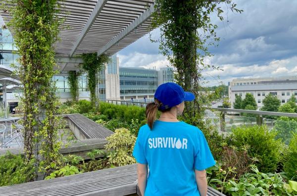 Elle Charnisky wears a ponytail and a blue cap with 'Survivor' printed on the back of her t-shirt stand in a rooftop garden overlooking Duke campus.
