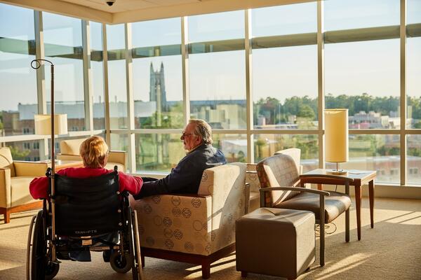 A woman in a wheelchair with her hsband in front of a window overlooking DCI buildings and greenery.