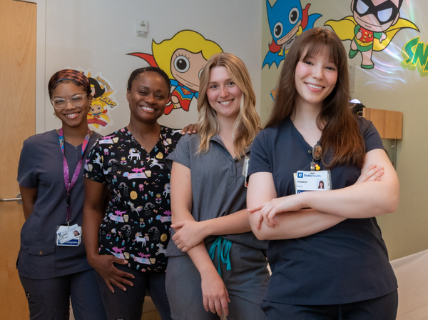 Four Duke Health imaging technologists stand together in a pediatric imaging room. Cartoon characters are pictured on the wall in the background.