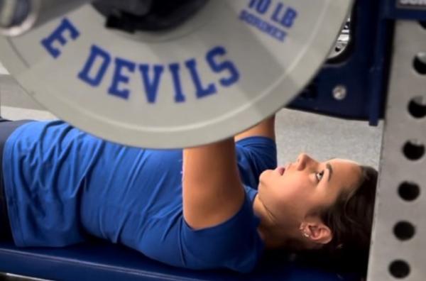 woman lying down bench pressing a large weight with the words "Devils"