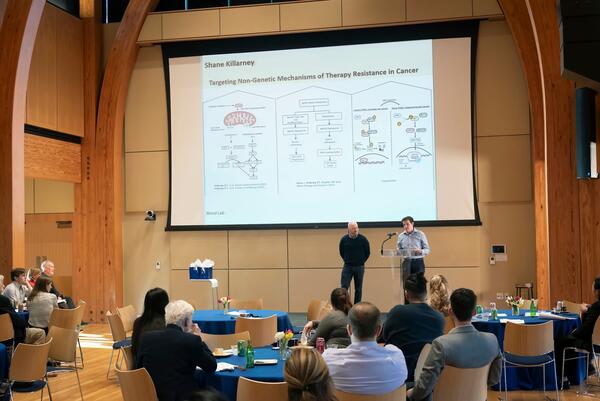 A young man and his mentor on stage in front of a screen showing a scientific presentation
