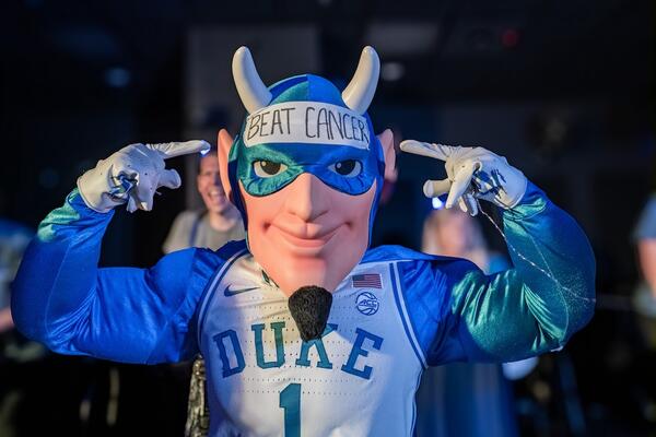 The Duke Blue Devil Mascot wears a Duke basketball jersey and a cape and points to a sign on his forehead that reads "Beat Cancer"