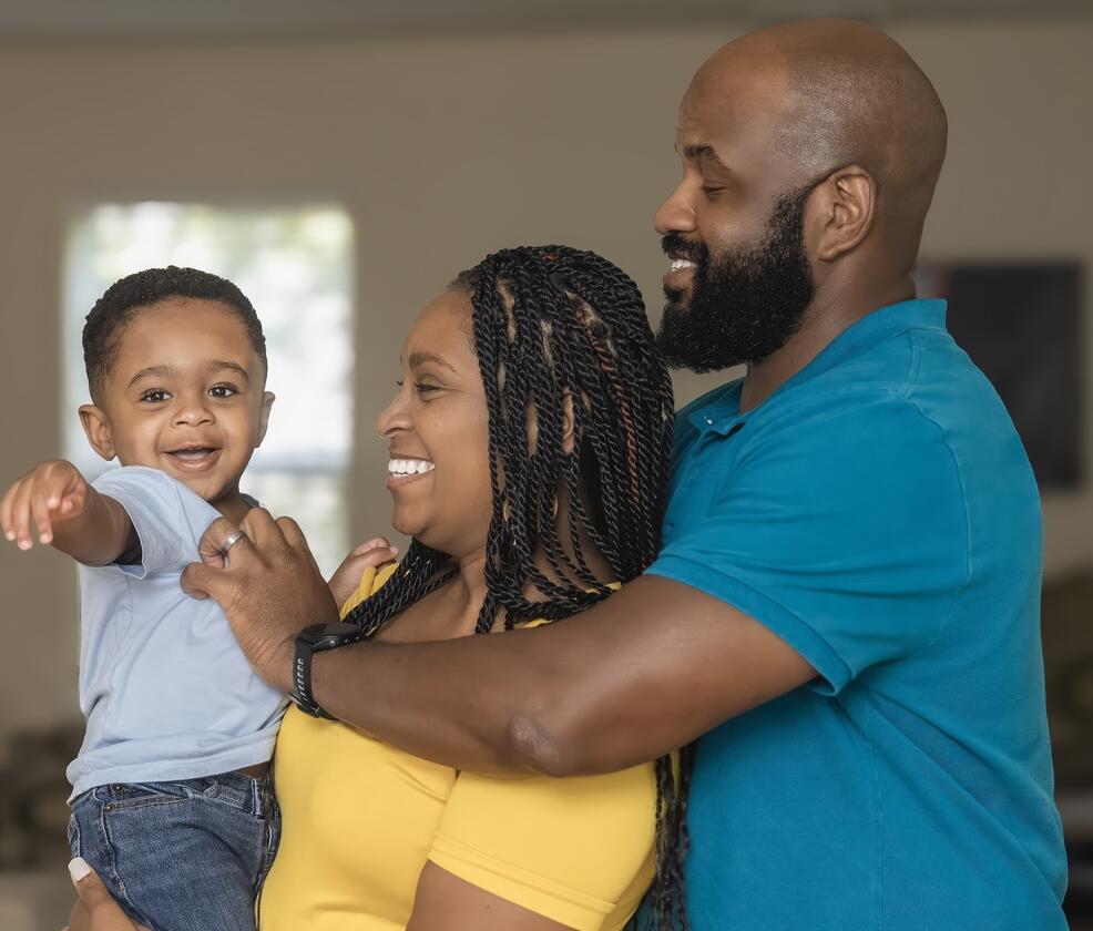 Two parents, one holding a toddler, smiling together as the toddler reaches toward the camera.