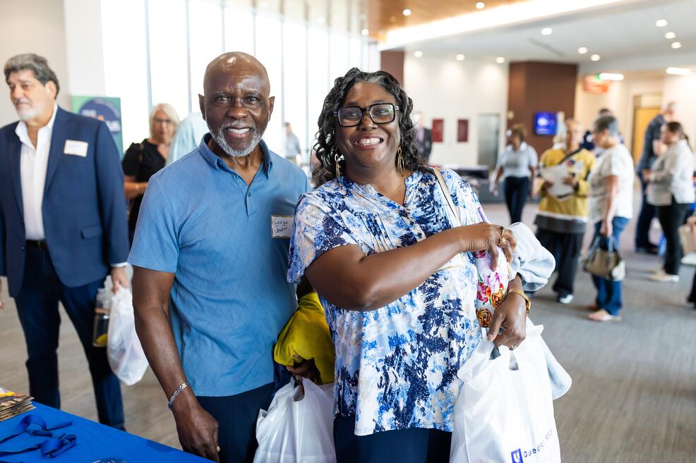 Black man with gre beard and black woman holding white bag with Duke CAncer Institute logo. They are standing in a large gallery filled with people