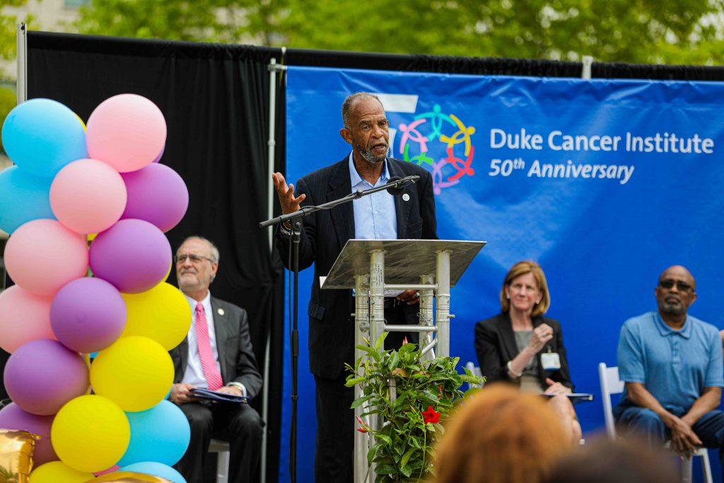 John Sanders gestures at the podium on stage and speaks. Michael Kastan, Mary Klotman, and Ovester Grays are seated behind him. The DCI 50th Anniversary Banner is the backdrop.