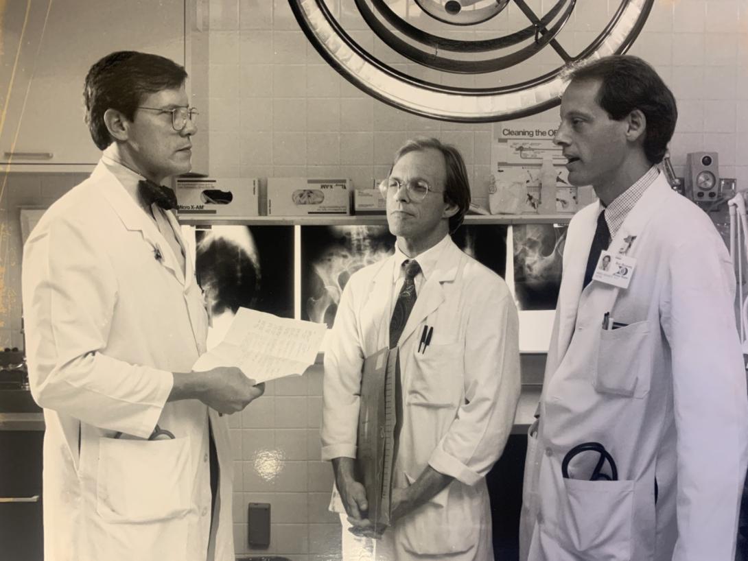 Three men in labcoats and ties confer with each other in the operating room.