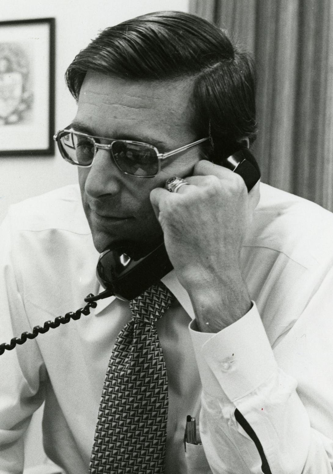 A man with glasses and a shirt and tie holds 70s era phone to his mouth and ear.