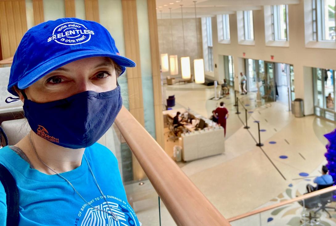 Elle Charnisky in blue cap with the word "relentless" has a black mask on and blue t-shirt. The backdrop is the lobby of Duke Cancer Center.