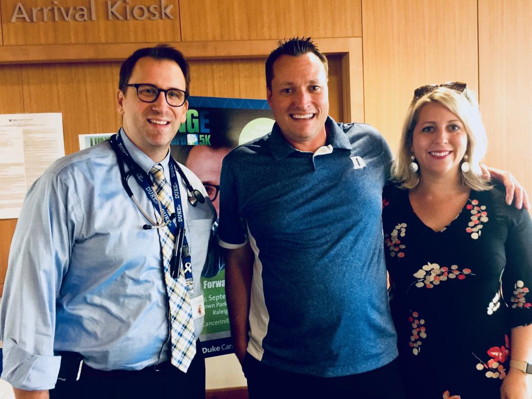 In the DCI thoracic clinic lobby, Jeffrey Clarke in a blue shirt, tie, and glasses, stands with Chad Eddy who's wearing a Duke shirt and his wife Missy who's dressed in a black flowered dress. 