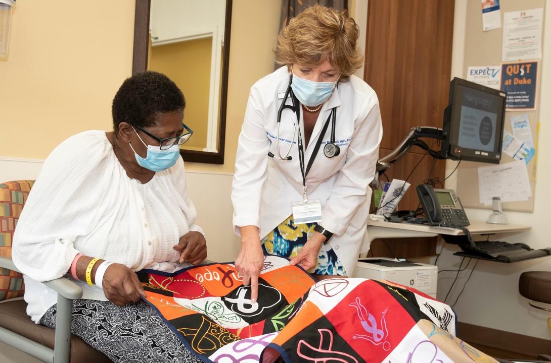 Henrietta Carr, in a mask, white top, and black print leggings, holds an orange, black, and white quilt. Susan Blackwell, PA, in a labcoat points to the white ribbon for lung cancer awareness.