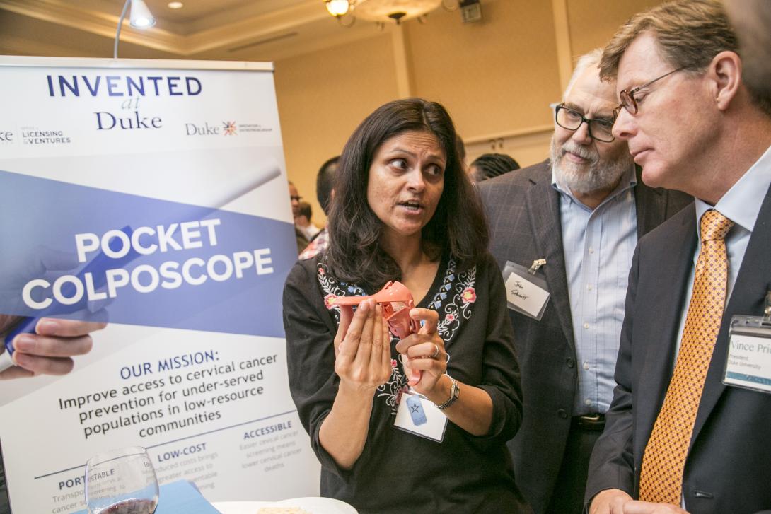 On a poster is printed "Invented at Duke" and "Pocket Colposcope." Nimmi Ramanujam, PhD, holds a pocket colposcope and shows it to two men in suits.