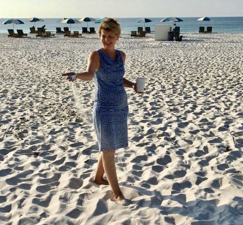 Tara Wilkes, in a blue dress, is at the beach smiling and watching her handful of sand slip through her fingers.
