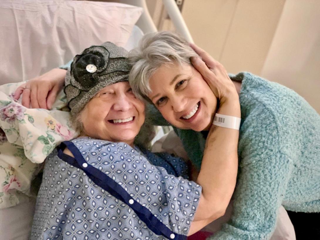Kathy Jennings hugs her friend Rebecca Gordon, who is wearing a hospital gown and a hat and sitting on a hospital bed.