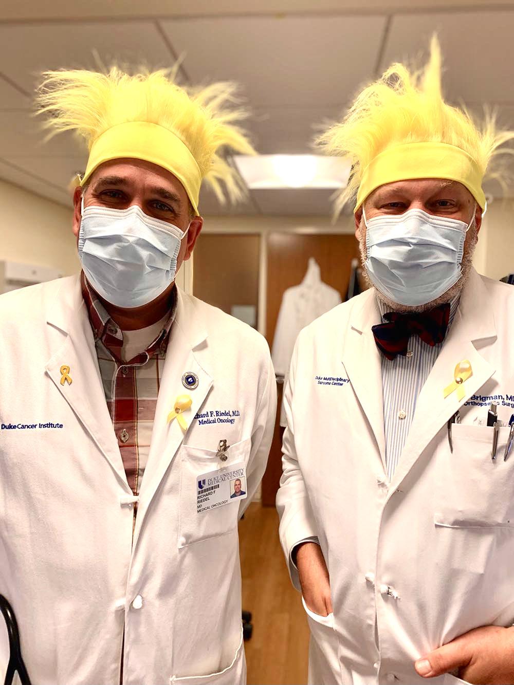 Two men in white coats and surgical masks with blond wigs