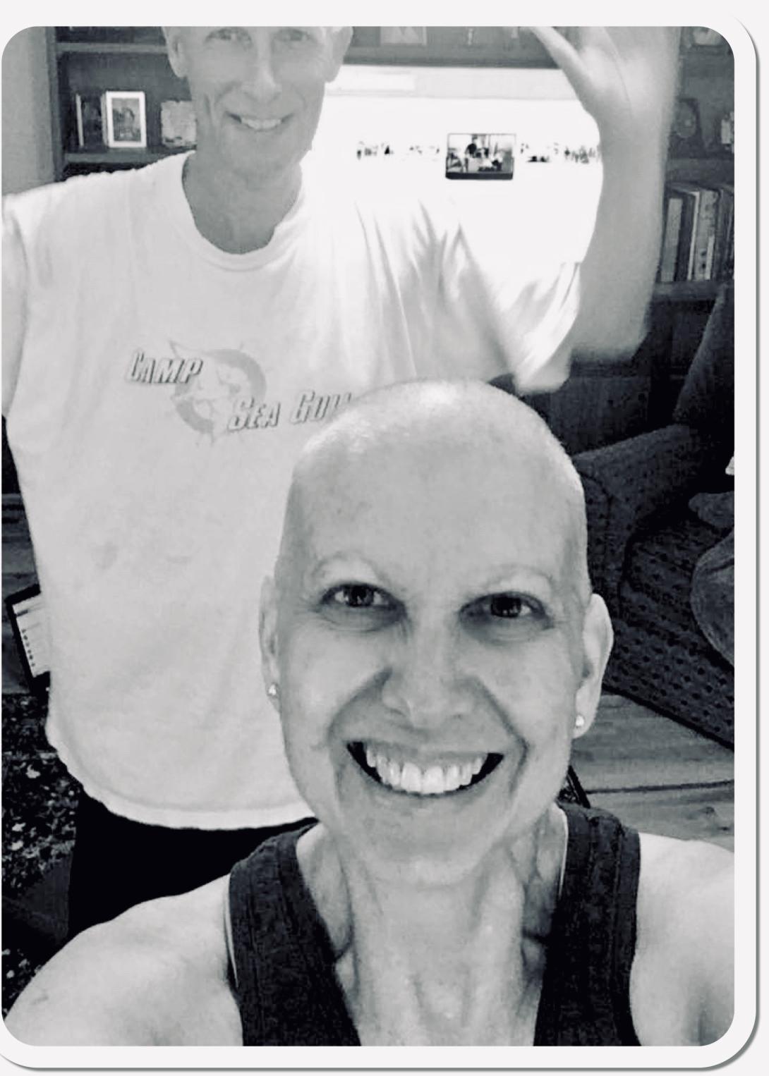 In this black and white photo, a bald Kathy Jennings smiles in a selfie with her husband in the background.