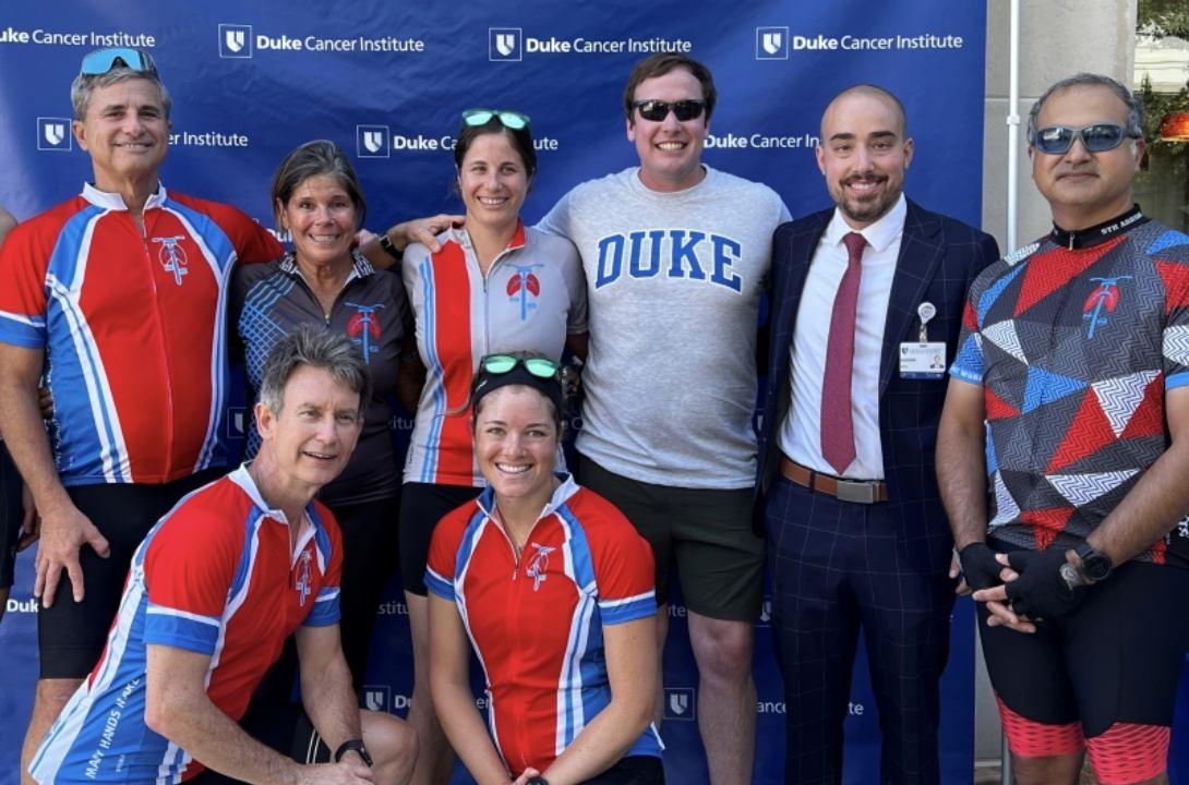 A group of cyclists stand and kneel together for a group picture in front of a Duke photo background.