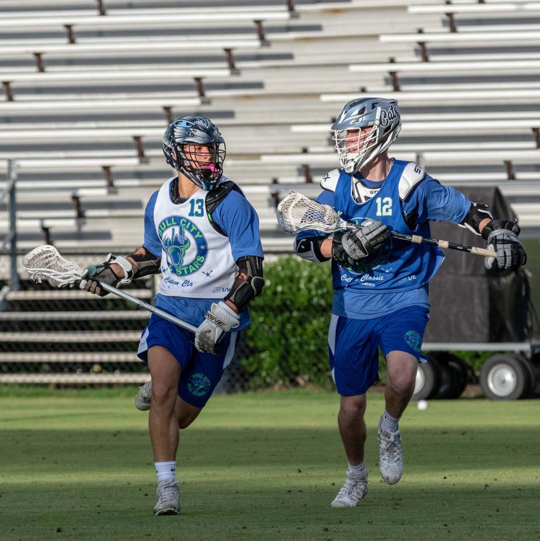 Two young men run in a lacrosse game. Both are wearing helmets and carrying lacrosse sticks.