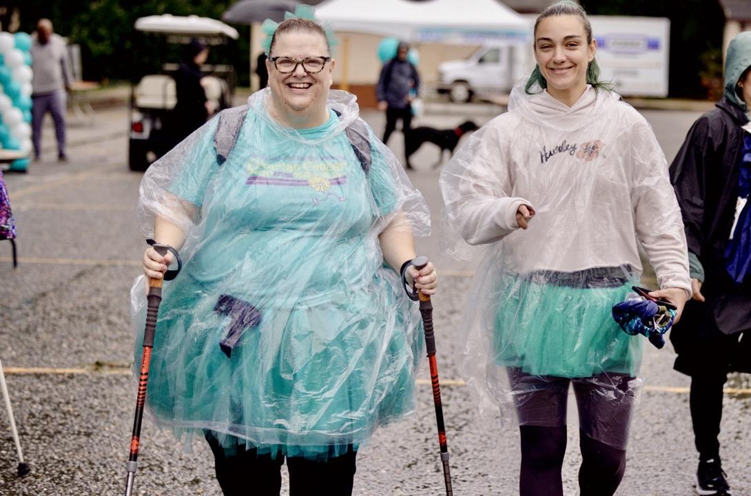A woman dressed all in teal with a clear rain poncho smiles. She walks with two canes and with a smiling female teenager beside her.