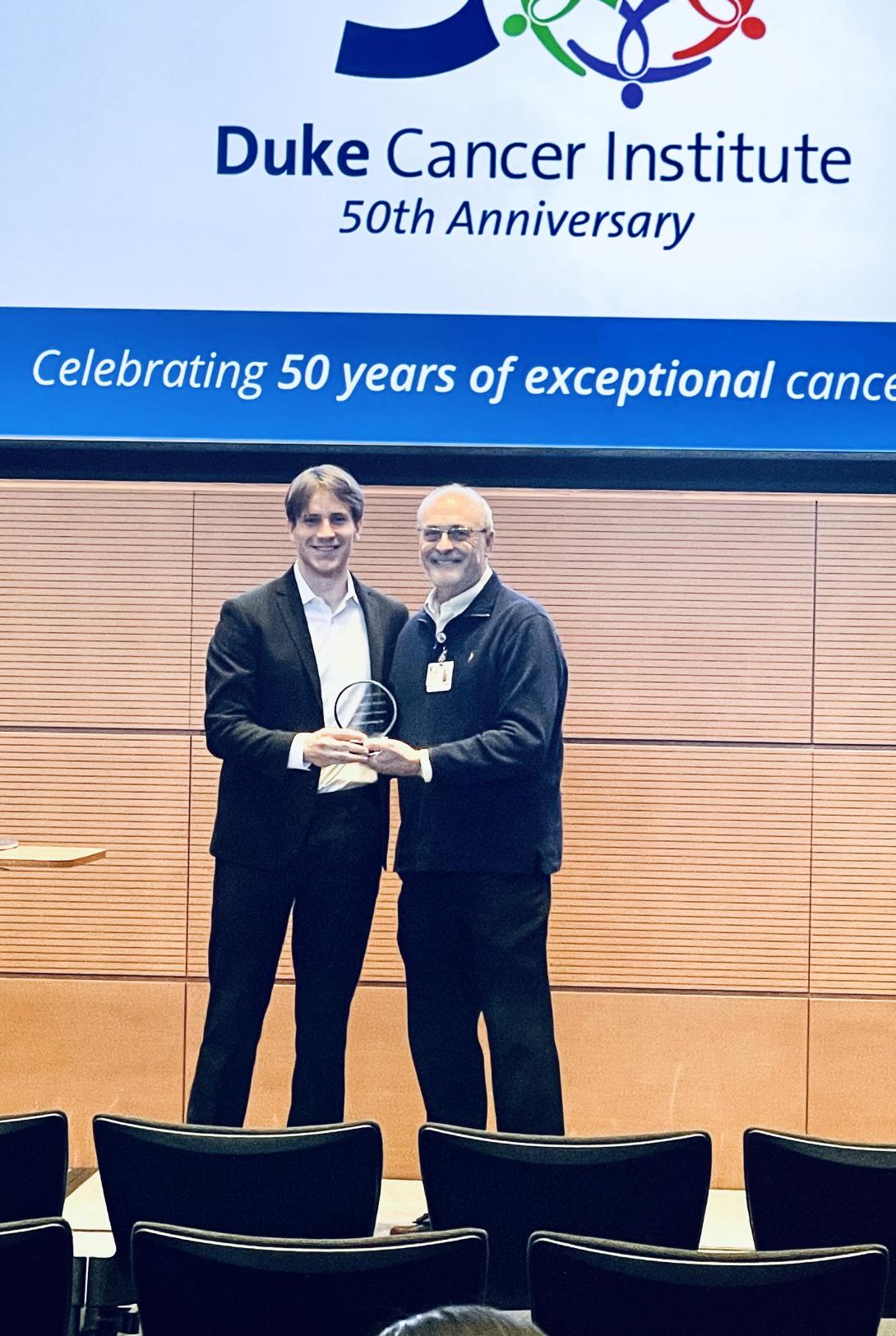 Two men hold a circular glass award in front of a screen with the Duke Cancer Institute 50th Anniversary logo and the words "Celebrating 50 yers of exceptional cancer care."