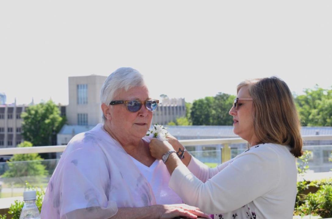 one woman pins a corsage on another woman outdoors with building in the background